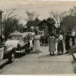 Three cars parked in a row with crowds of students standing around on the street. A sign visible on one of the cars says, Augsburg College. The photo is for the sendoff of a car caravan taking wheat for India to Washington, D.C. in May 1951.