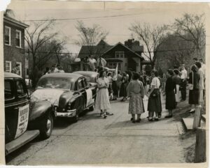 Three cars parked in a row with crowds of students standing around on the street. A sign visible on one of the cars says, Augsburg College. The photo is for the sendoff of a car caravan taking wheat for India to Washington, D.C. in May 1951.