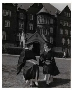 two women in cap and gown sit on bench in front of Old Main