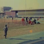 A runner running on the track and a small group sitting and standing on the Stadium field, circa 1970