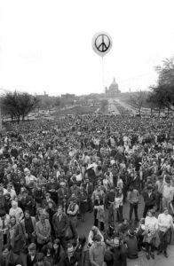 Huge crowd of peope amassed at the Minnesota state capitol, Spring 1970. The Cathedral is seen in the far background, and a balloon with a peace sign floats in the sky