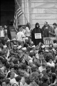 Crowd of people on the steps of the Minnesota state capitol in Spring 1970; a person in a black shroud dressed as Death has a sign that says, "Death Wins All Wars"