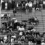 Spectators, many holding umbrellas, in the bleacher seats at MIAC track meet, Spring 1969