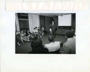 Mayor Cisneros standing at the front of the Fine Arts Lounge speaking to groups of students seated around him in October 1985