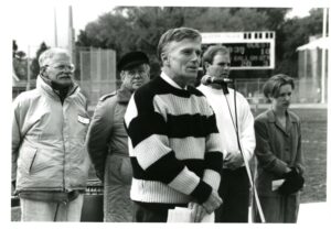 Image of President Gavin speaking at a microphone on the athletic field, with four other people behind him.
