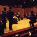 Students in a semi-circle on stage performing in African Music Ensemble Spring 1994