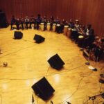 Students in a semi-circle on stage performing in African Music Ensemble Spring 1994