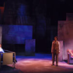 An image from the 2007 production Angels in America. A character laying in a bed with another character standing beside the bed. On the other side there is a character sitting on a couch and another character standing next to the couch.