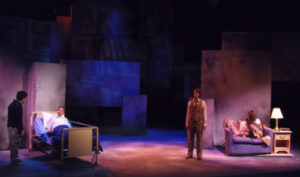An image from the 2007 production Angels in America. A character laying in a bed with another character standing beside the bed. On the other side there is a character sitting on a couch and another character standing next to the couch.