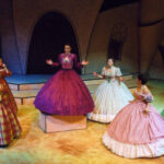 An image from the 2007 production of Lysistrata. There are seven ladies in fancy dresses, six of them are staring at the same character who is on a pedestal.