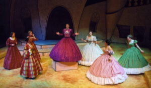 An image from the 2007 production of Lysistrata. There are seven ladies in fancy dresses, six of them are staring at the same character who is on a pedestal.