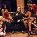 An image from the 2008 production the Threepenny Opera. A guy character is surrounded by six women on a couch
