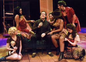An image from the 2008 production the Threepenny Opera. A guy character is surrounded by six women on a couch