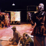 An image from the 2008 production of the Threepenny Opera. Half the characters are in a group, far away from the camera looking at the other half, which are next to the camera.
