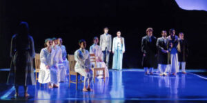 An image from the 2009 production of Our Town. A group of female characters are sitting on chairs and a group of guys are standing a distance away. Behind them there is a guy and girl standing together.