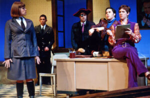 An image from the 2010 production of the Accidental Death of an Anarchist. Three characters are on a desk and an other character is standing a distance from the desk.