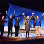 An image from the 2010 production of the Accidental Death of an Anarchist. Seven characters are standing in a line facing the audience, six of them have both arms in the air.