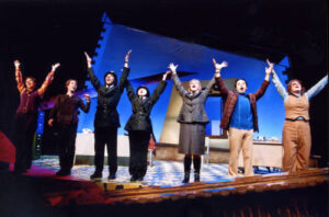 An image from the 2010 production of the Accidental Death of an Anarchist. Seven characters are standing in a line facing the audience, six of them have both arms in the air.