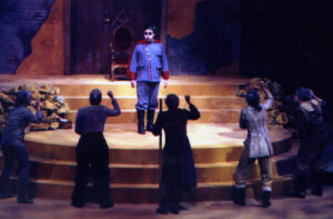 An image from the 2011 production of Antigone. Five characters are standing below the platform waving a fist at a character standing on the platform